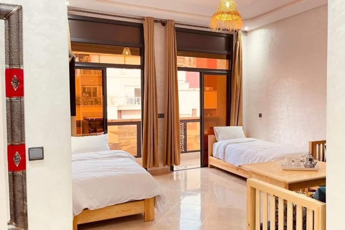 A hotel room with two beds, a wooden table, and a view through large windows.