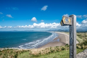Ein hölzernes Wegweiser mit einem kreisförmigen Emblem und Richtungspfeilen, mit Blick auf einen Strand mit Wellen und entfernten Bergen.