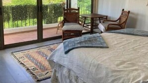 Ein Schlafzimmer mit einem großen Bett, Blick auf Grün durch das Fenster und ein Balkon mit Stühlen.