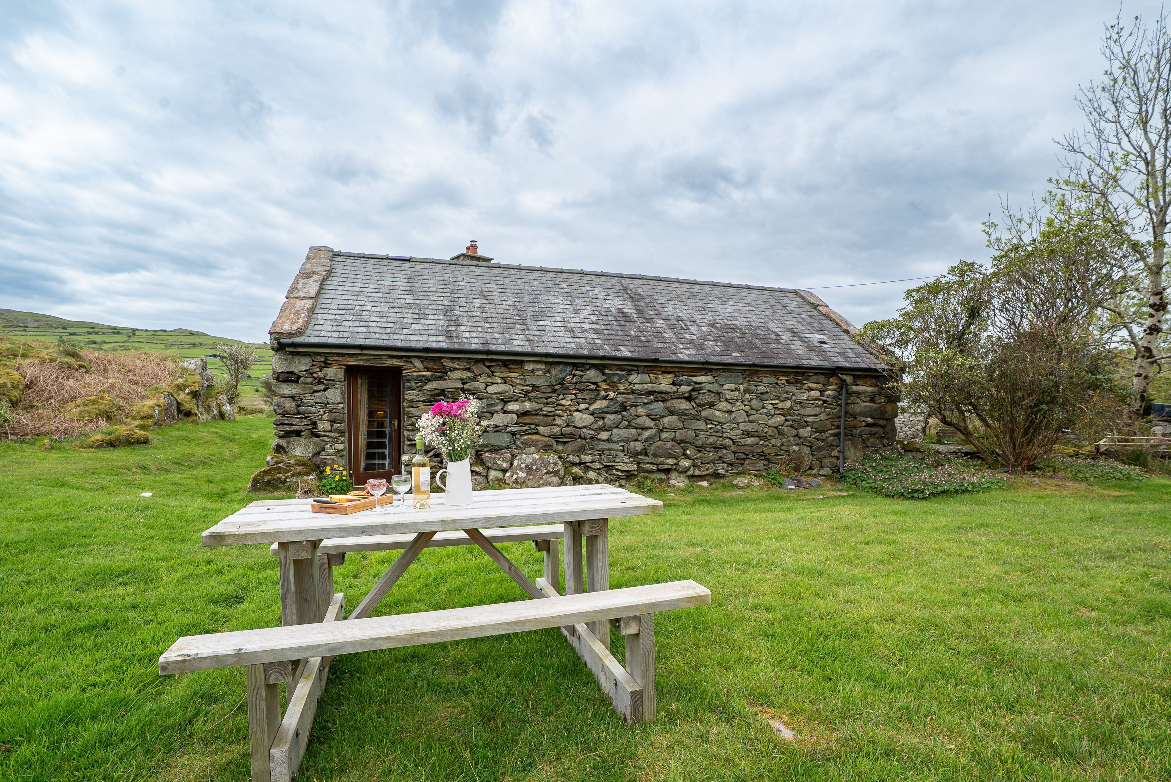 A stone cottage with a wooden table and benches in a grassy yard.