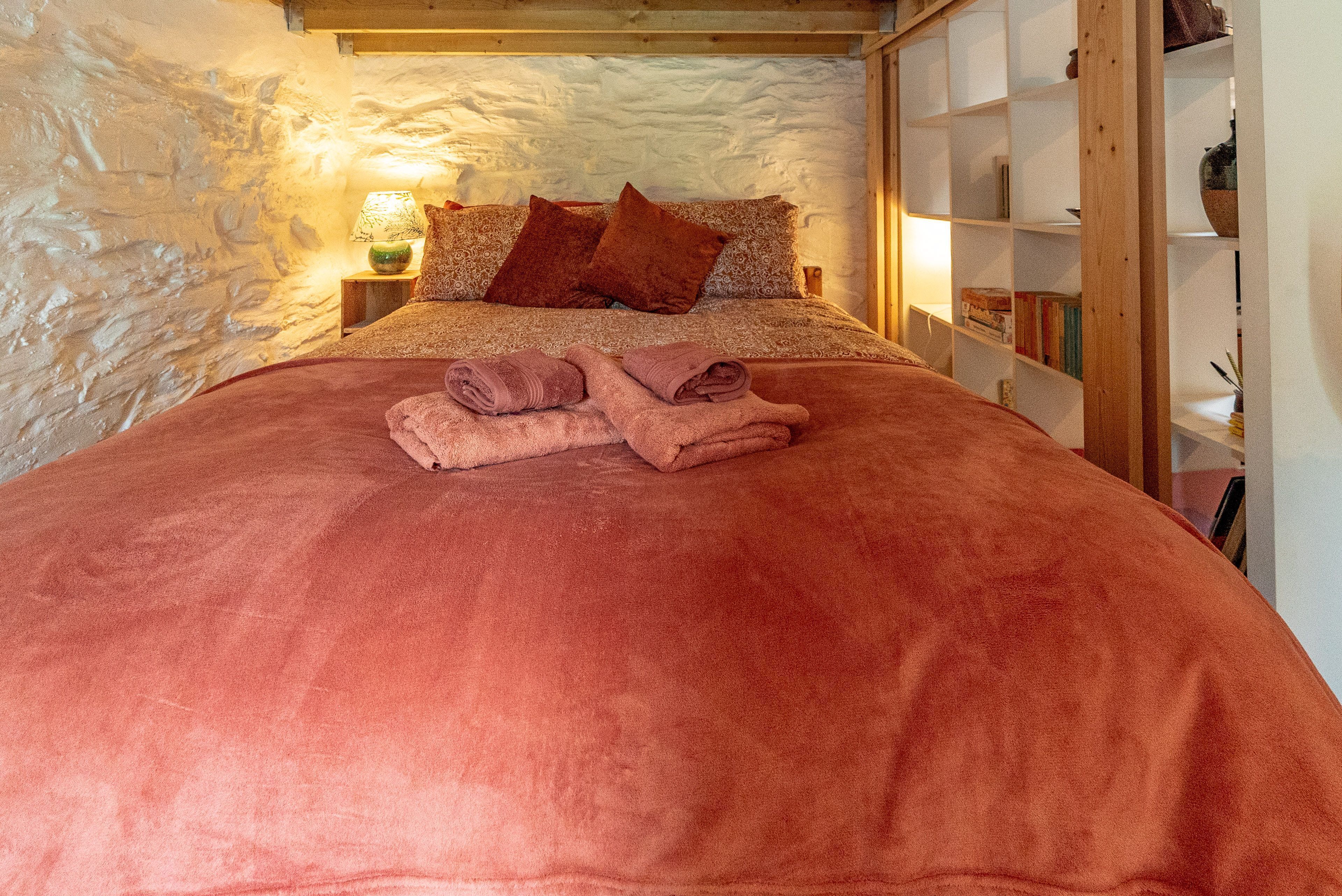 A bed with a red blanket, pillows, and folded towels. There is a nightstand with a lamp and a shelf with books and a vase.