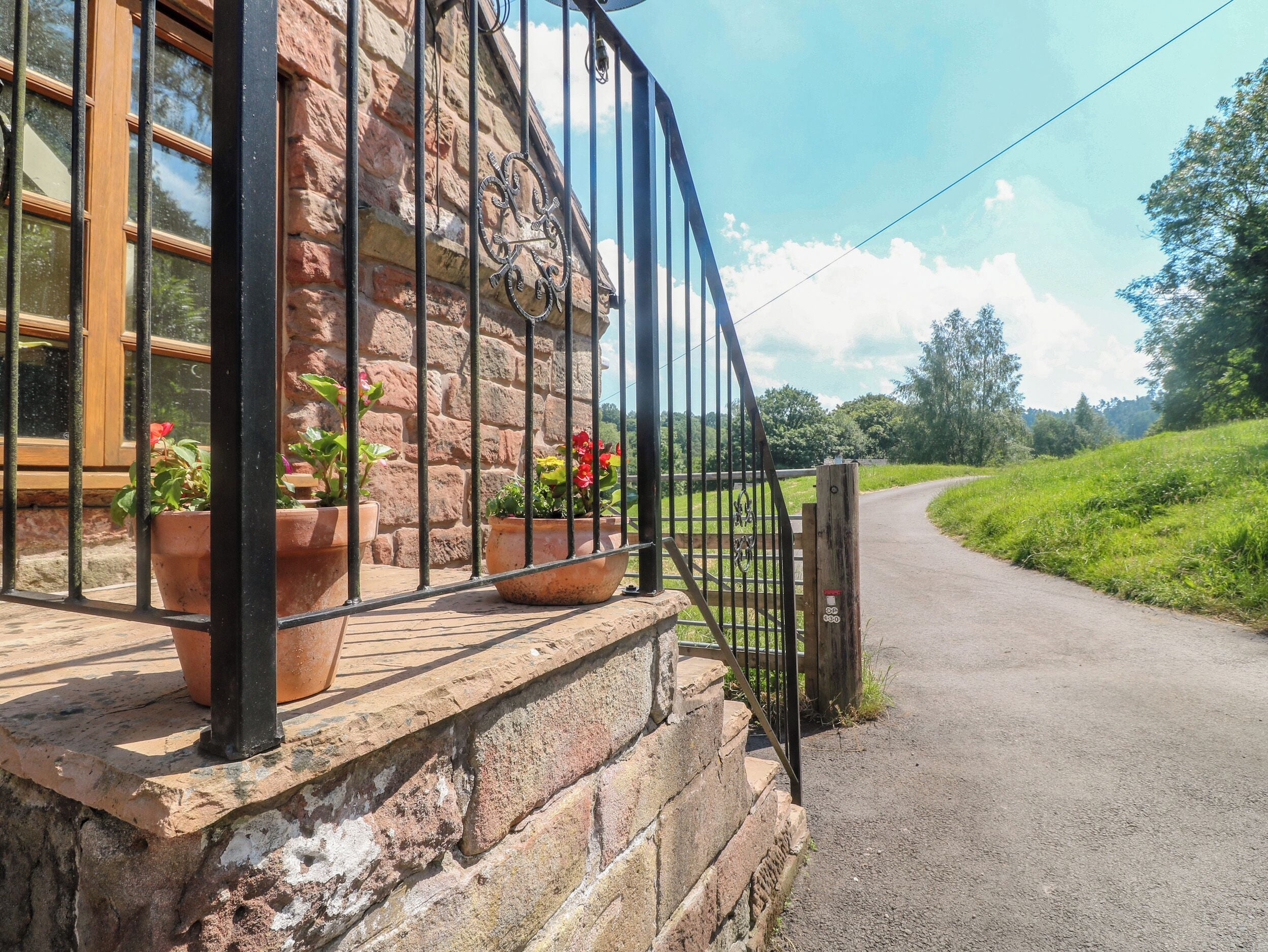 A stone building with a metal gate, potted plants, and a road leading into a green landscape.