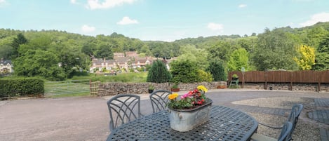 A patio with a round table and chairs, a flower pot, and a view of a village and hills in the background.