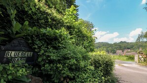 Un camino rural con un cartel que dice 'BLUEBELL BARN' rodeado de vegetación densa y un cielo despejado.