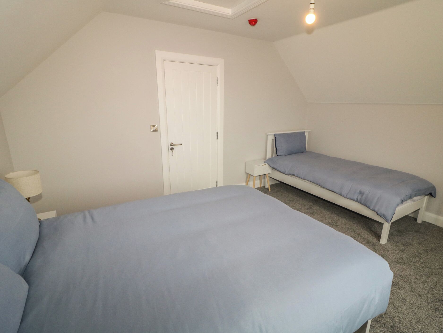 A room with two beds, a white door, a small bedside table, and a ceiling light.
