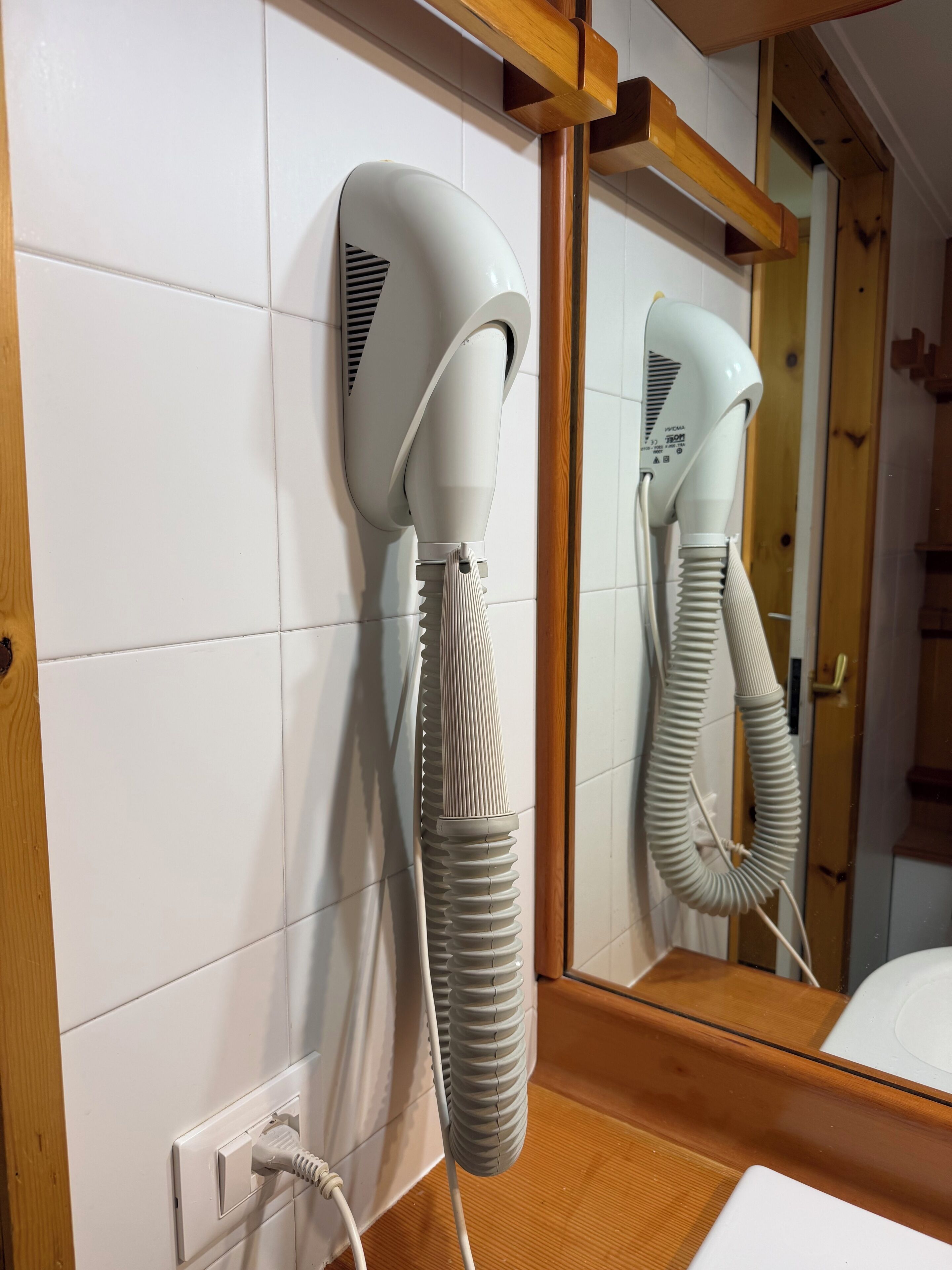 Un sèche-cheveux fixé sur un mur recouvert de carreaux blancs, une étagère en bois au-dessus, et un miroir qui reflète la pièce.