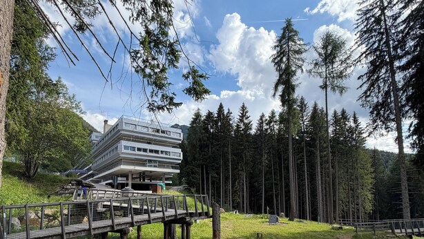 A multi-story building with a balcony, surrounded by a forested area with a wooden bridge and picnic tables.
