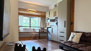 A modern room with a wooden desk, a bed with a patterned bedspread, a kitchenette with cabinets, and a view of a garden through a window.