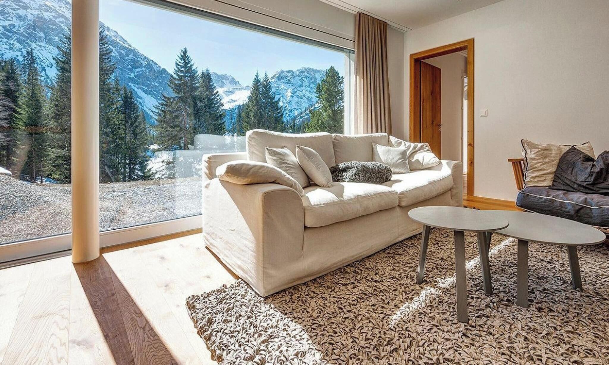 Ein modernes Wohnzimmer mit einer Couch, zwei Beistelltischen und Blick auf schneebedeckte Berge durch große Fenster.