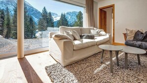 A modern living room with a sofa, two side tables, and a view of snow-capped mountains through large windows. - Fürggli Appartement/Fewo, Dusche und Bad, WC, Erdgeschoß/Parterre by Interhome (Arosa)