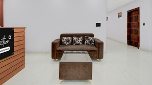 A reception area with a wooden desk, a brown sofa with patterned cushions, and a glass-top coffee table. - Premium Budha Tower (Noida)