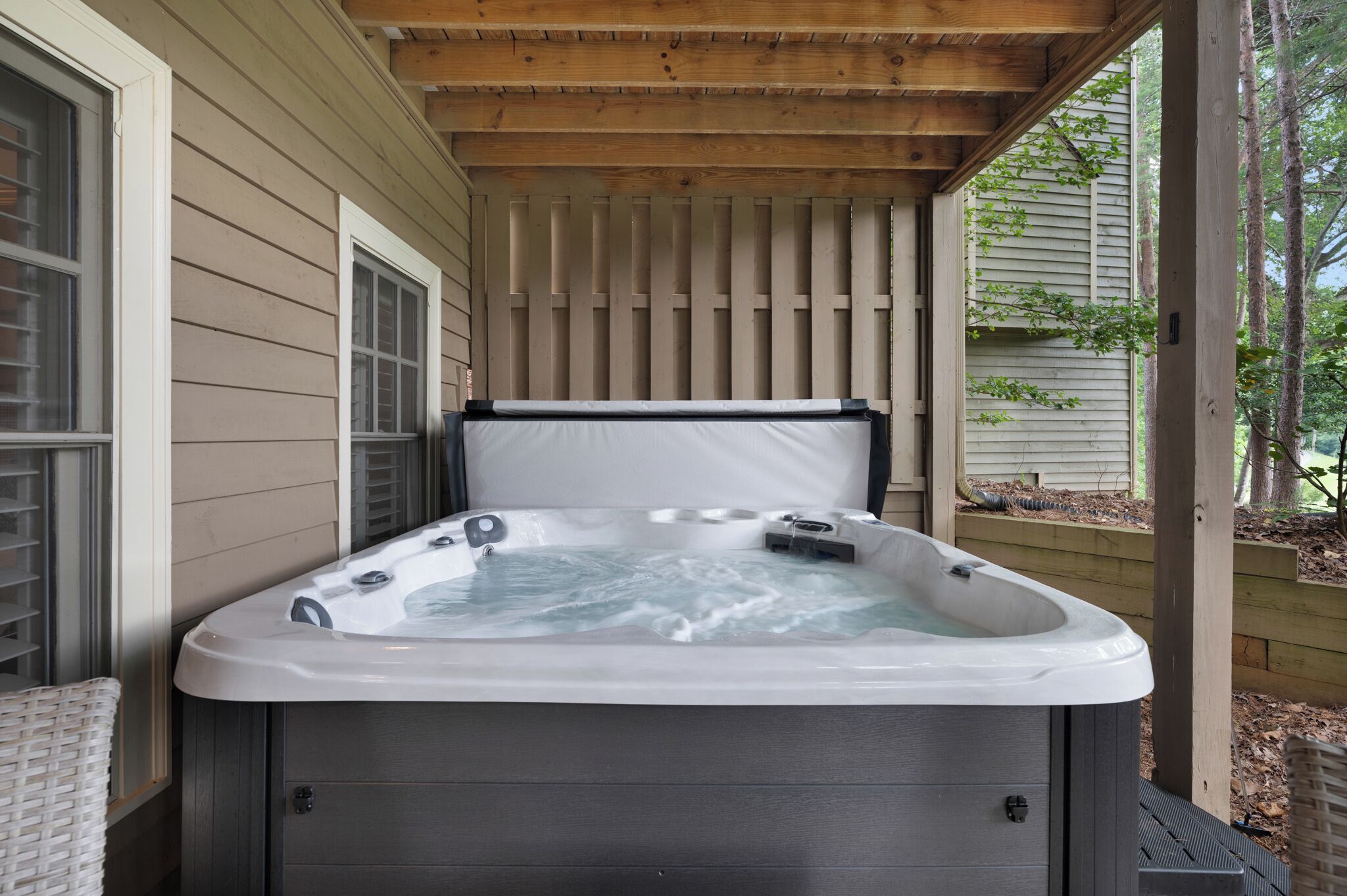 A hot tub under a wooden pergola with a view of a house and trees.