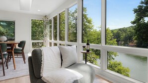 Ein Zimmer mit einem großen Fenster mit Seeblick, ein graues Sofa mit weißer Decke und ein Essbereich mit Stühlen und einem Tisch.