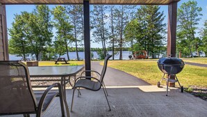Uma área externa coberta com mesa, cadeiras, churrasqueira e vista para um lago.