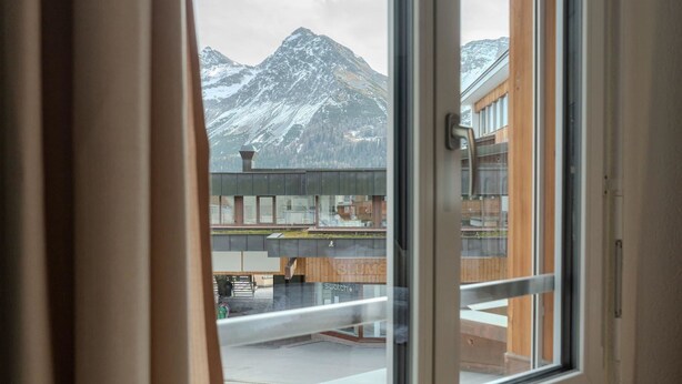 Une vue à travers une fenêtre, avec une montagne en arrière-plan, un bâtiment portant une enseigne avec le mot « Swatch », et une structure en bois au premier plan.