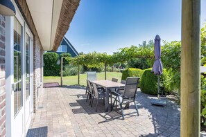 A patio with a table and chairs, a thatched roof, and a view of a garden. - Lavish 4-person Beach Villa in Makkum (Makkum)