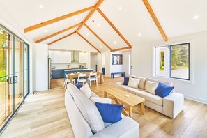 A spacious living area with a high ceiling, wooden beams, a sofa set, a dining table, and a kitchen in the background. - Beach House w/ Patio & Game Room (CoastalC) (Long Beach)