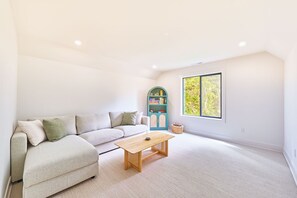 Ein geräumiges Wohnzimmer mit einem hellen Sofa, einem Holztisch und einem Fenster mit Blick auf Bäume.