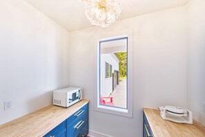A room with a microwave on a wooden counter, a small table with a device, and a window with a view of a building and trees. - Beach House w/ Patio & Game Room (CoastalC) (Long Beach)