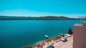 Paesaggio costiero con cielo sereno, acqua, colline in lontananza e un edificio con un balcone che si affaccia sulla scena.