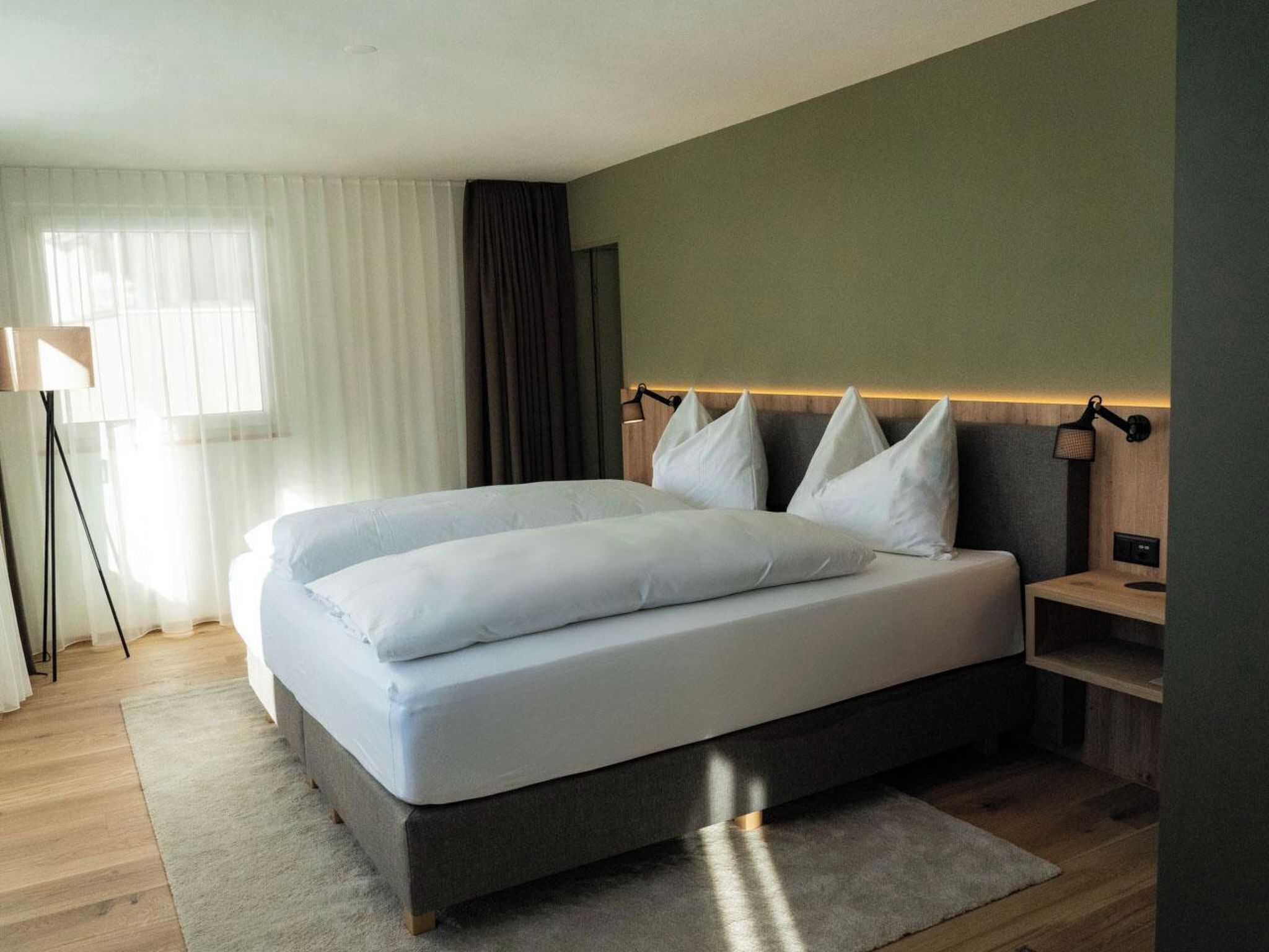 Una habitación de hotel moderna con una cama grande, una mesita de noche, una lámpara y una ventana con cortinas.