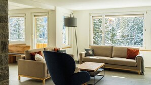 Ein modernes Wohnzimmer mit Sofa, Sessel und Couchtisch, große Fenster mit Blick auf schneebedeckte Bäume und einer Steinwand.