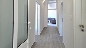 Un pasillo con puertas correderas, suelo de tonos claros y vistas a una sala de estar con sofá y ventanal.