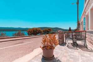 Un espace extérieur aménagé avec une terrasse dallée, des tables et des chaises, offrant une vue sur un plan d’eau et des collines.