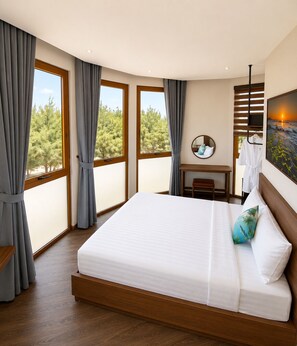 A hotel room with a large bed, a desk, a chair, a mirror, and a view of trees through the windows.