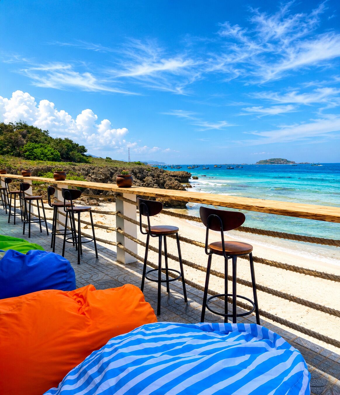 Eine Strandbar mit Außenbereich, inklusive Stühle und Sitzsäcke, mit Blick auf das klare blaue Meer und eine felsige Küste.