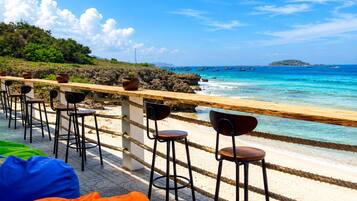 Eine Strandbar mit Außenbereich, inklusive Stühle und Sitzsäcke, mit Blick auf das klare blaue Meer und eine felsige Küste.