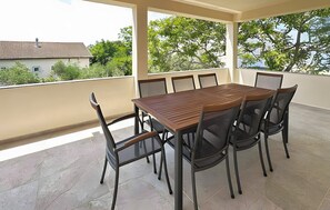 Un patio techado al aire libre con mesa y sillas de madera, suelo de baldosas y vistas a edificios y árboles.
