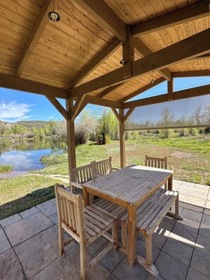 Un espacio al aire libre cubierto con una mesa y bancos de madera, con vistas a un lago y vegetación.