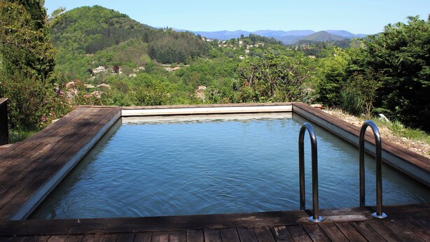 Una terraza de madera que conduce a una piscina rectangular con barandillas de metal, rodeada de exuberante vegetación y montañas al fondo.