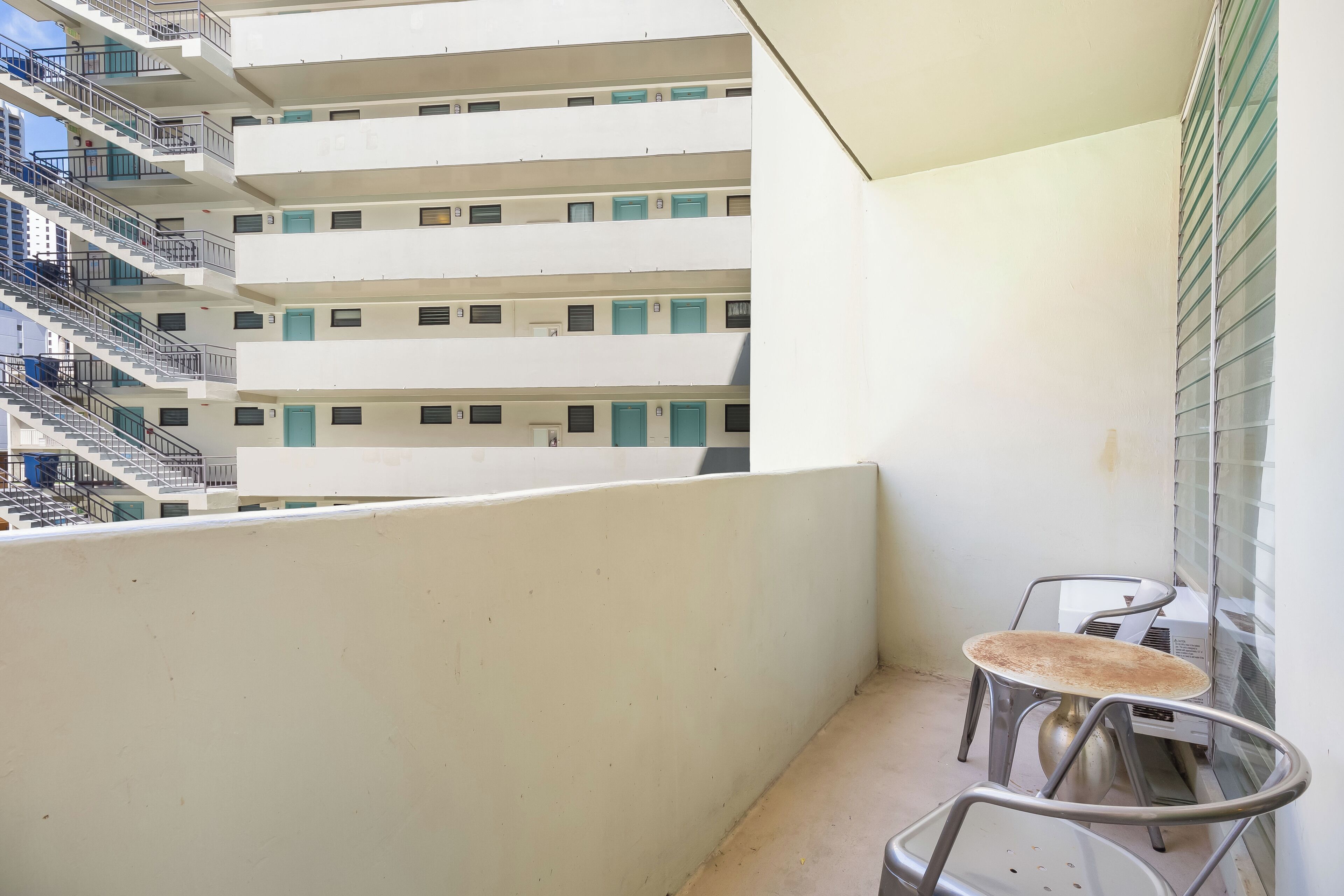 A balcony with a table and chairs, a window with blinds, and a building with multiple floors and balconies in the background.