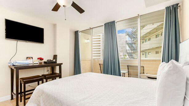 A hotel room with a bed, a desk with a laptop, a television, and a view of a building through the window. - Chic Studio w/Kitchenette, Lanai, AC, WiFi–Waikīkī Grand #501 (Honolulu)