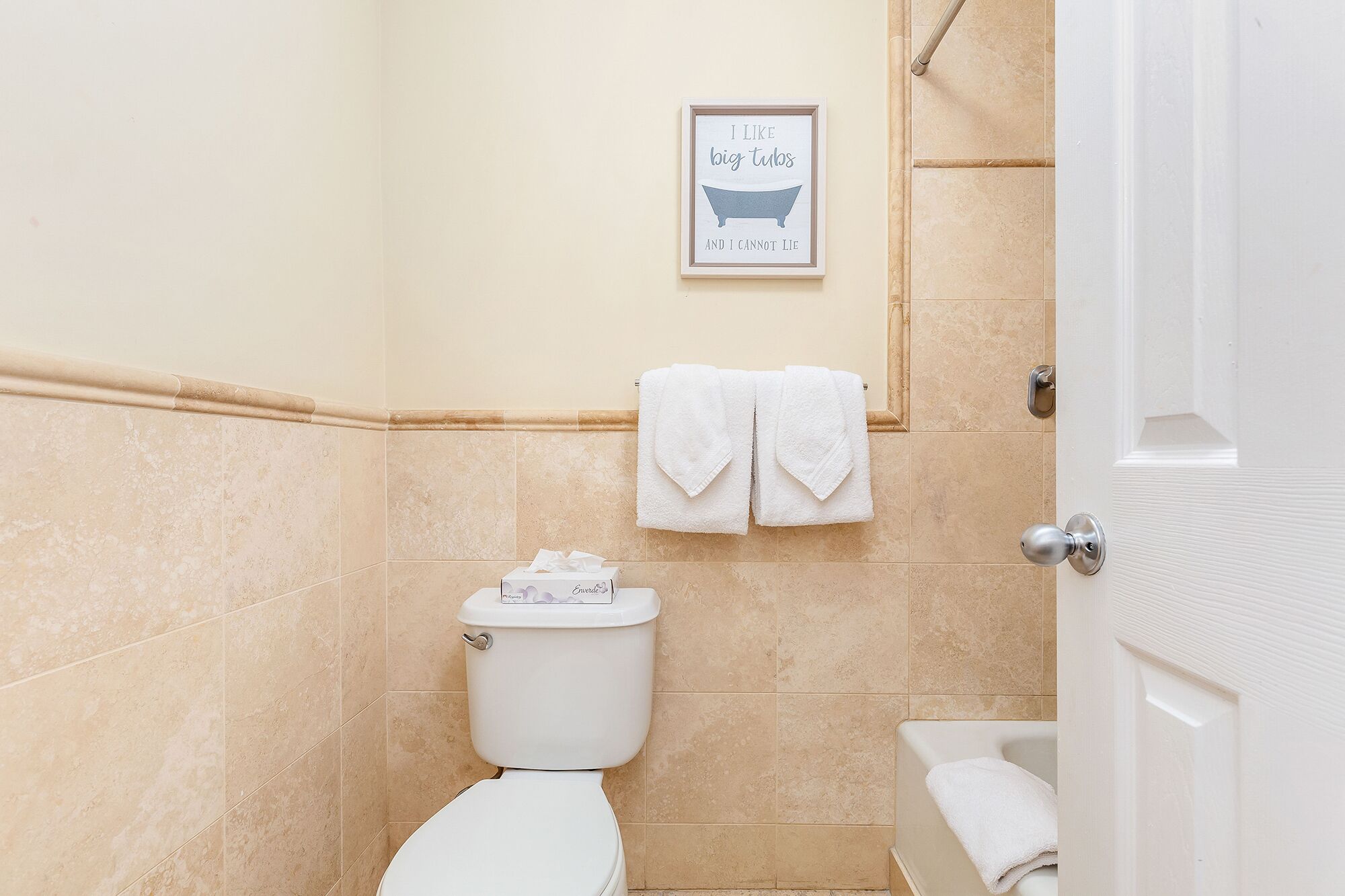 Un baño con baldosas color crema, un inodoro blanco, una cita enmarcada en la pared y una bañera con una toalla.