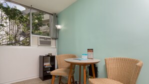 A small room with a window, a round table, two wicker chairs, a bookshelf, and a wall-mounted air conditioner. - High-Style Studio w/Kitchenette, WiFi, Flat Screen+AC–Waikiki Grand 503 (Honolulu)