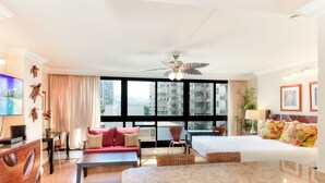 Habitación de hotel con cama, mesa de comedor, sofá, televisor y ventana grande con vista a la ciudad.