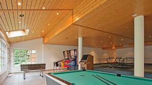 Ein Billardtisch mit Spielstöcken, ein Fußballtisch und eine Dartscheibe in einem Raum mit Holzbalkendecken und großen Fenstern.