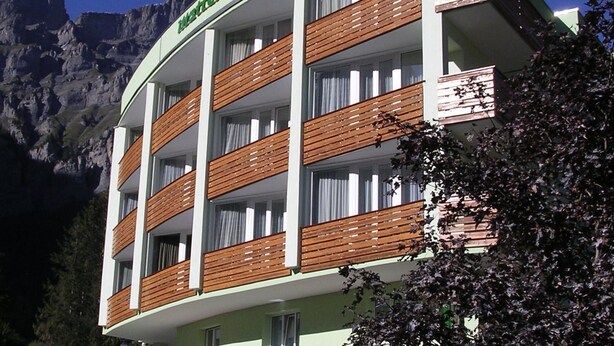 Un hotel de varios pisos con balcones, un paisaje montañoso de fondo y un letrero que indica que es un hotel.