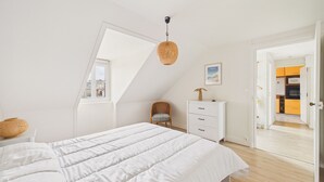 Un dormitorio con una cama grande, una silla, un aparador y una puerta que da a una cocina.