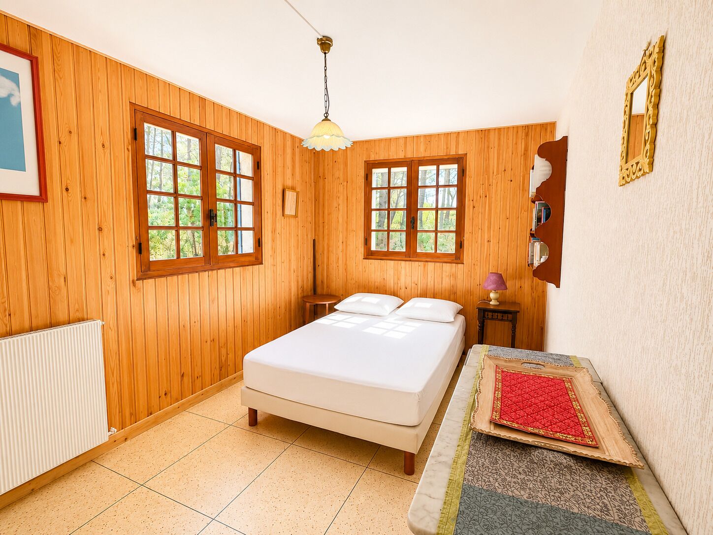 A wooden-paneled room with a bed, two windows, a radiator, a lamp, and a small table with a quilted table runner.