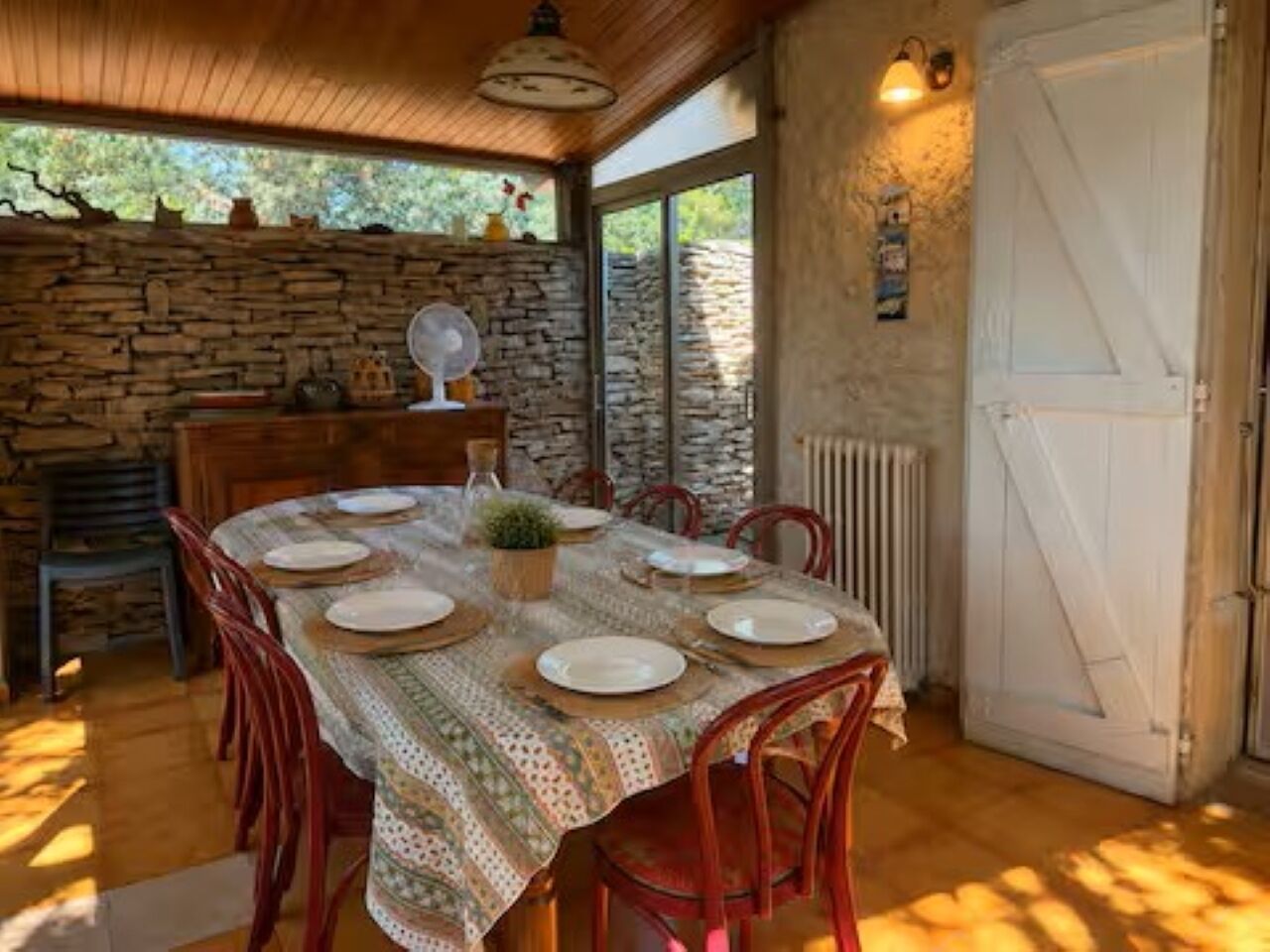 Un coin repas avec une table ronde dressée pour quatre personnes, entourée de chaises en bois, d’un mur en pierre et d’une porte blanche.