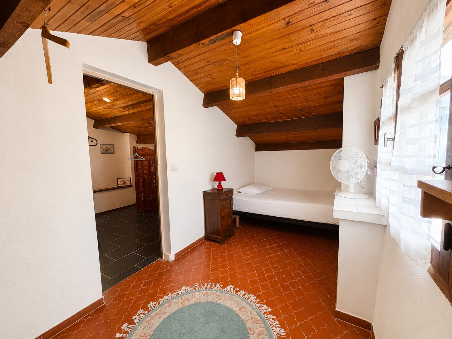A bedroom with a wooden ceiling, a bed, a fan, a red lamp, and a small table.