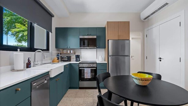 A modern kitchen with teal cabinets, a stainless steel refrigerator, a black dining table with a bowl, and a window with a view of trees. - Cozy 2BR in Miramar | Kitchen, Laundry & TVs (San Juan)