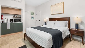 Quarto de hotel com uma cama, mesas de cabeceira, um pequeno espaço de cozinha e um quadro na parede.