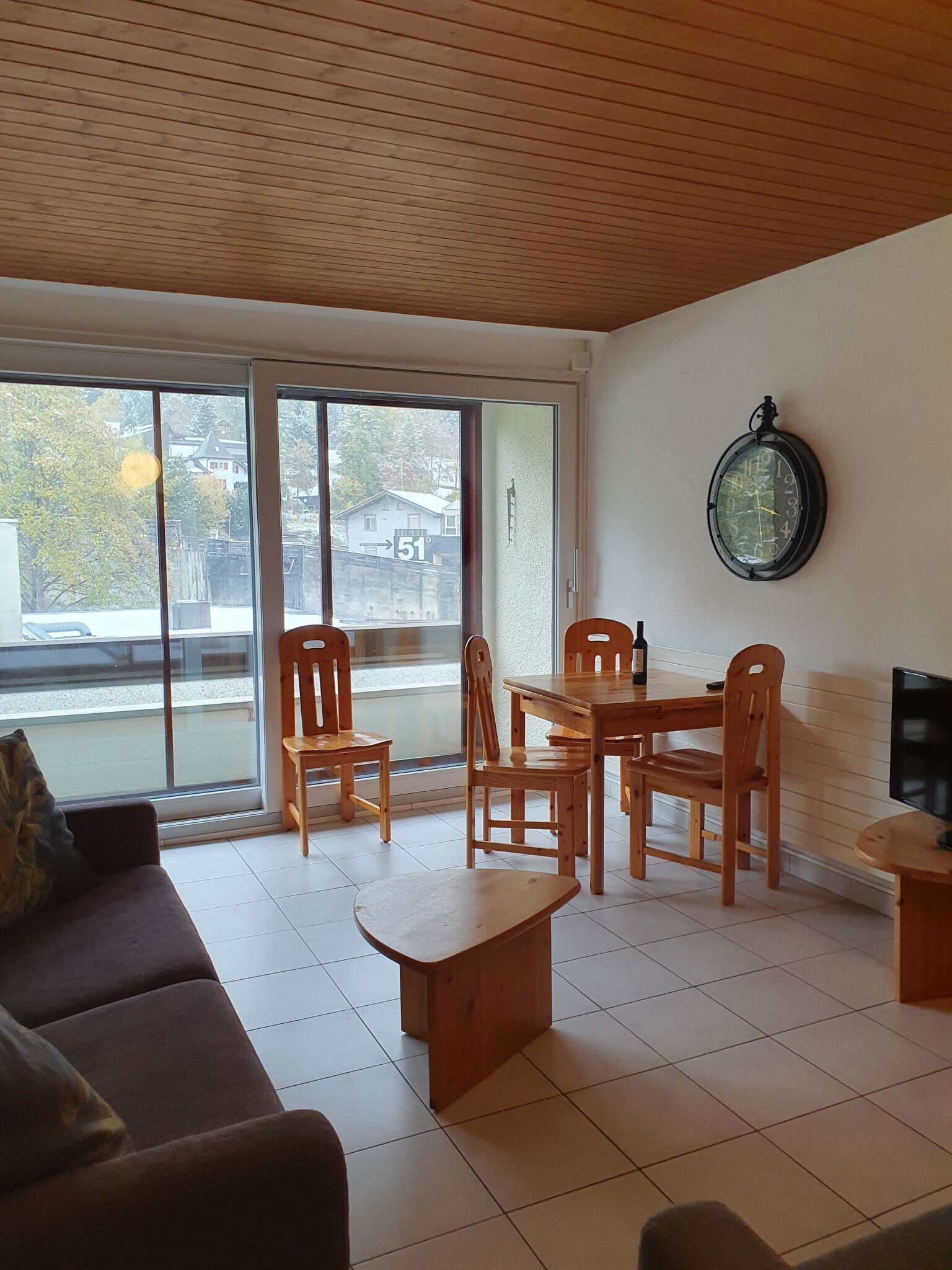 Una sala de estar con sofá, mesa de centro y mesa de comedor con sillas. Hay un televisor en un mueble bajo y un reloj en la pared.