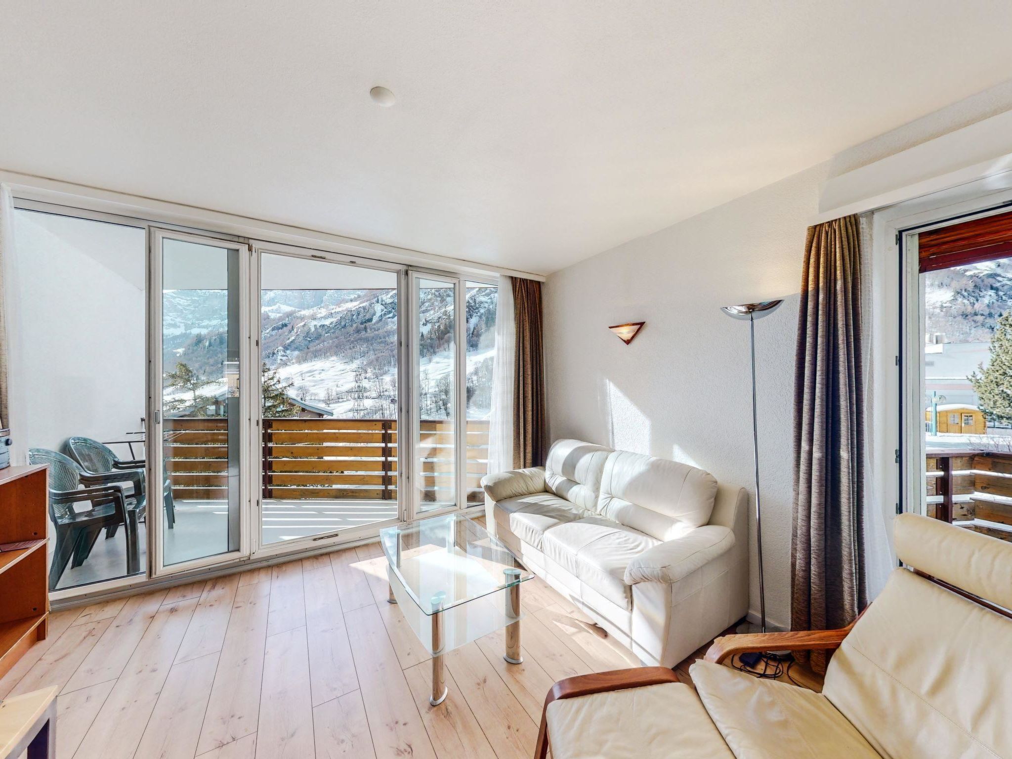 A modern living room with a glass sliding door leading to a balcony, a white sofa, a glass coffee table, and a wooden floor.