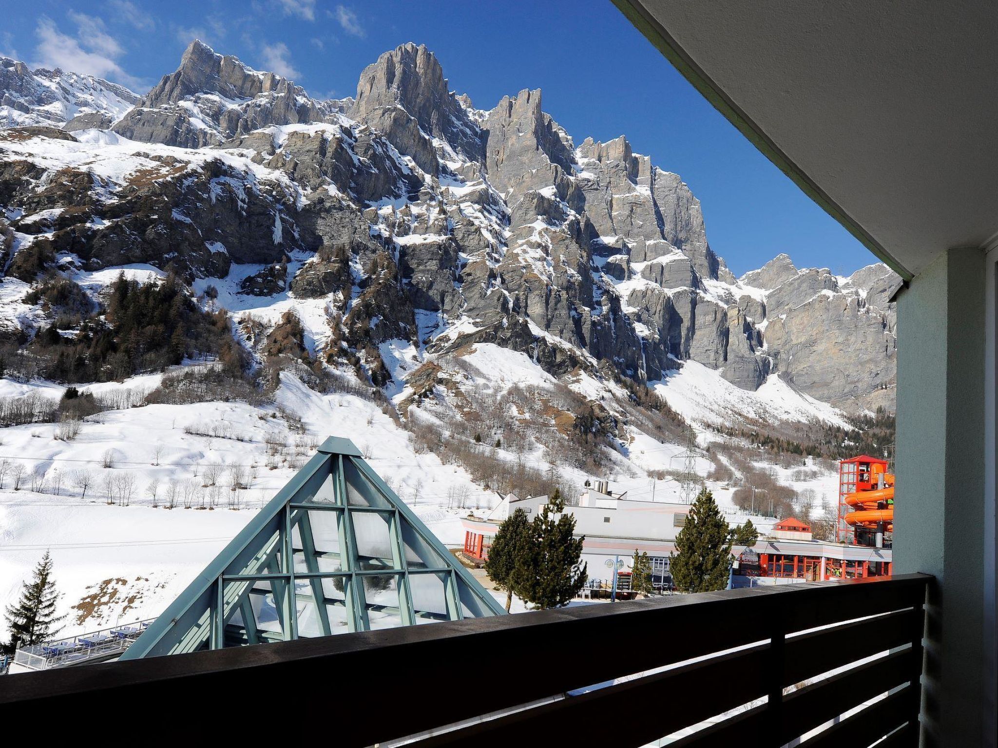 A mountain view from a balcony with snow-covered peaks, a glass structure, and a red building in the foreground.